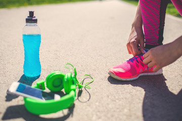Young Fitness girl tying shoes before start running. Female runner with energy drink and headphones on the track. Sport recreation concept. Healthy lifestyle.