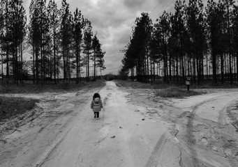 Little girl walking towards a crossroads on a rural dirt road in the country with pine trees in black and white