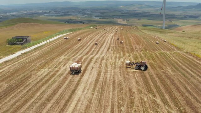 Aerial shot of a hay tractor collecting and loading hay bales on a trailer in a beautiful landscape of Basilicata in Southern Italy with wind turbines in the background generating green energy