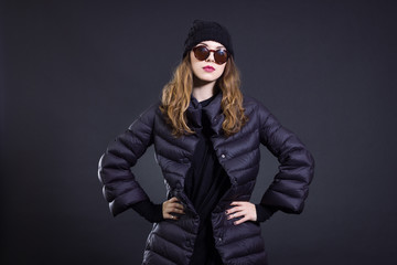 A stylish young woman in a black down coat and knitted hat poses on a black background. Autumn and winter women's fashion.