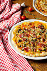 Fresh homemade pizza with prosciutto, capers, olives on a wooden background in composition with a red cloth and olive oil. Italian Cuisine. Selective focus on center of pizza