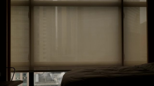 Automatic sunscreen curtain slowly move up, empty bed in small room of hotel. Modern city seen outdoors through full height window