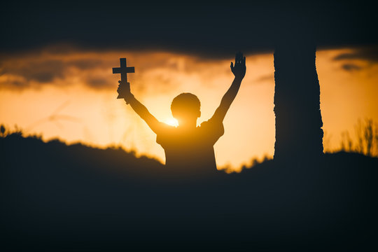 Young man sitting and lift hands for worshipping God with cross at sunset background. christian silhouette concept.