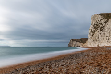 Fototapeta na wymiar Long exposure of waves crashing into the beach and white chalk cliffs on a cloudy day in England, UK.