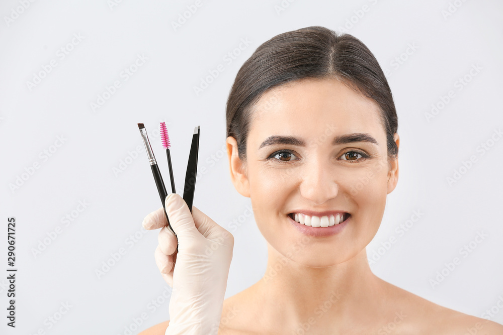 Wall mural young woman with beautiful eyebrows and hand of cosmetologist holding tools against light background