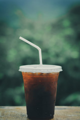 Ice coffee (espresso, cappuccino, latte) in plastic cup and white staw on wooden table, vintage tone with blurry green nature with copy space for your text. Vacation and relaxing concept.