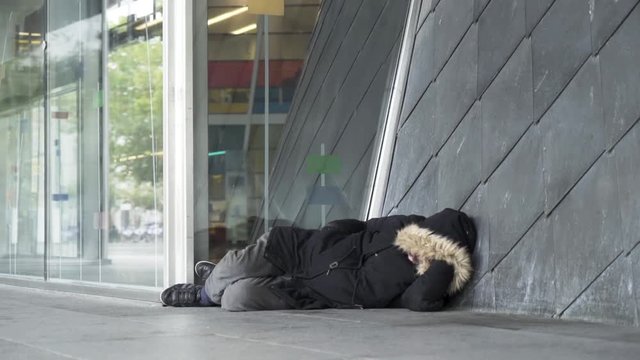 Homeless man in warm clothes sleeping outdoors near the entrance of the building, poverty concept. Stock footage. Poor unemployed male tramp lying near the concrete wall.