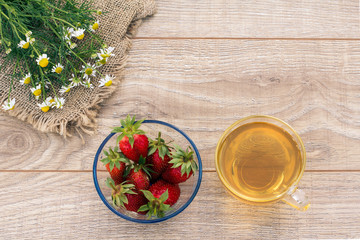 Glass cup of green tea, stawberries with white chamomile flowers on wooden background.