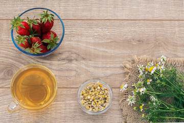 Glass cup of green tea, stawberries with white chamomile flowers on wooden background.