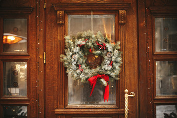Stylish christmas wreath with red bow and berries at front doors of store at holiday market in city street. Space for text. Rustic decoration. Christmas street decor. © sonyachny