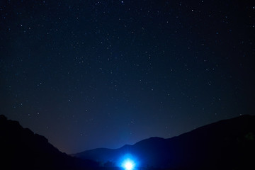 Night starry sky over the mountains. Stars and constellations are visible through light clouds. The bright light of the headlights of a car in the distance.