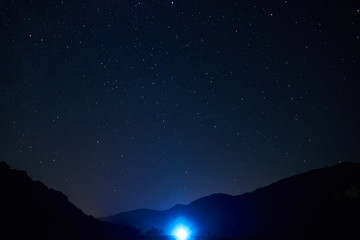 Fototapeta premium Nocne niebo gwiaździste nad górami. Gwiazdy i konstelacje są widoczne przez lekkie chmury. Jasne światło reflektorów samochodu w oddali.