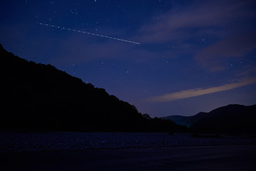 Meteor flies in the night starry sky over the valley of a mountain river.
