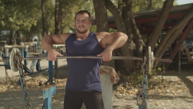 Muscular male bodybuilder working out in outdoor gym doing exercises with barbell. Athletic fit man performing upright -row weigth training exercise, lifting barbell straight up to collarbone outdoors