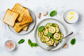 Grilled zucchini salad with basil leaves, yogurt sauce and fried bread in a simple ceramic plate on a white concrete background. Flat lay with copy space.