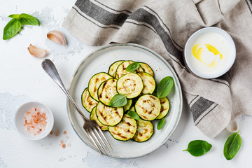 Grilled zucchini salad with basil leaves, yogurt sauce and fried bread in a simple ceramic plate on a white concrete background. Flat lay with copy space.