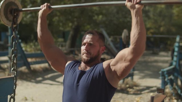 Muscular athletic man training shoulders, lifting barbell while sitting on bench during outdoor workout. Confident determined bodybuilder pumping up muscles, exercising with barbell in outdoor gym.