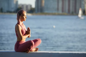 Fototapeten Lotusblume Young healthy woman sitting with eyes closed in lotus position and meditating in the city with sea in the background  © AnnaStills
