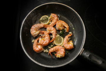 tiger prawn shrimps are roasted in a black pan with garlic, lemon and herbs on a black stove, high angle view from above