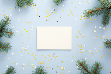 Blank paper greeting or invitation card on blue background with fir tree branches and glitter confetti stars. Christmas, New Year, winter holidays concept.