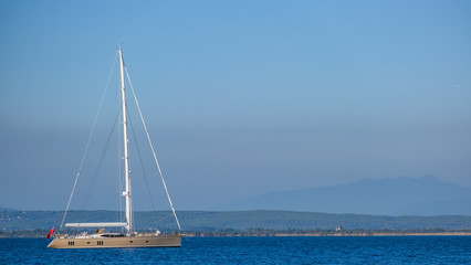 Fototapeta premium Single white gold sailboat on calm water in a sunny day in the Mediterranean on the background of the islands and blue sky. Water activities, vacation at sea, happy holidays.