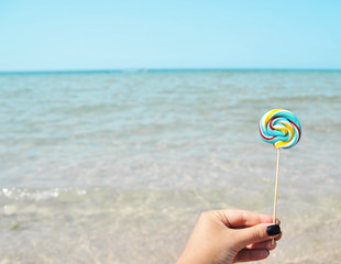 Female hand holds colorful candy on sea background.