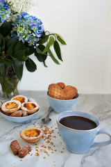 Cup of homemade chocolate accompanied by puff pastry cakes filled with custard and cookies. In the background a blue flowers.
