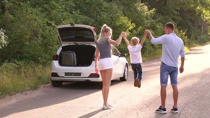 Young parents raise their young son holding hands on an empty road in the woods. A little boy jumps with his parents on the road near the car with trunk open. Family having fun during the trip by car.