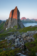 View from Mount Hesten on Iconic Mountain Segla in light of midnightsun in front of clear sky and mountain range in background, rocks and boulders in foreground, Fjordgard, Senja, Norway