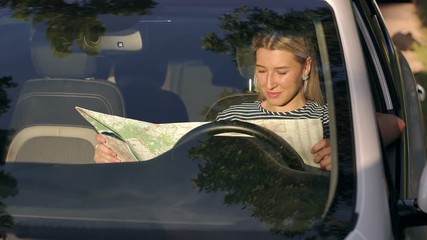 Young blonde girl studying a map of the area sitting behind the wheel of a car at sunset. The girl travels by car in the summer. Slow motion.
