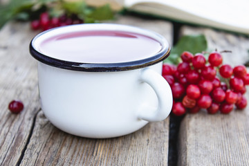 White mug or cup of hot viburnum tea on a wooden table near an open book and red viburnum berries. Source of natural vitamins. Used in folk medicine.