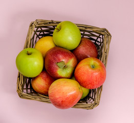 Ripe juicy apples of new crop. Group of colorful apples in a small wicker basket isolated on pink background. Symbol of healthy lifestyle. Fruits rich in vitamins, iron. Harvest festival, organic food