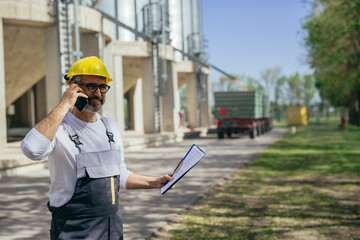worker using mobile phone standing in front of grain silos © cherryandbees