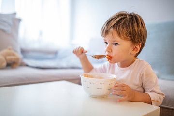 Eating cheerful baby boy with messy face. Portrait of a toddler eating. Open mouth child with an appetite eats millet porridge. Happy cute baby kid eating food itself with spoon.