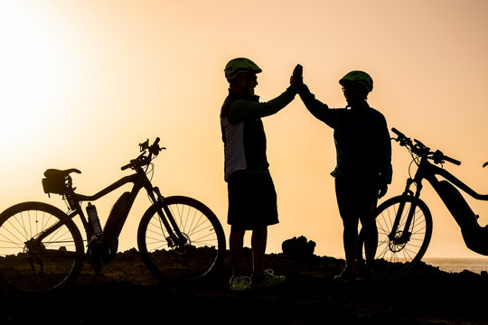 couple of two seniors and maturepeople together at the sunset with their bikes giving five with the silhouetteand the sea at the background