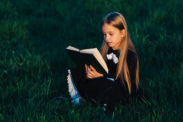 A little girl in black sits on the grass and holds a green book in the light of the setting sun. A...
