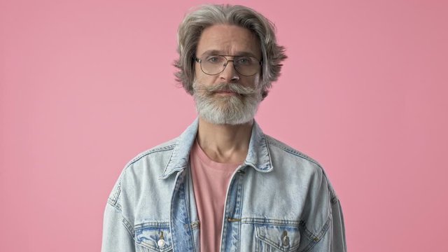 Displeased elderly stylish bearded man with gray hair in denim jacket disagree with someone and gesturing with hands while looking at the camera over pink background isolated