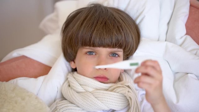 The face of a cute little boy with temperature lying at sofa home. Portrait of sick baby child little boy with high fever in bed. Cold flu virus disease, sick kid. 4 K