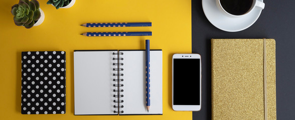 Obraz na płótnie Canvas Modern design of office desk with blank paper of organizer with mock up. Smartphone, cup of coffee and pencils with notebook on yellow and balck background. Panoramic flat lay.