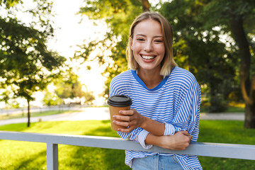 Image of smiling cute woman holding paper cup and bending over railing