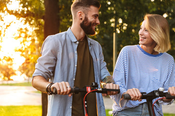Image of caucasian cheerful couple smiling while riding e-scooters