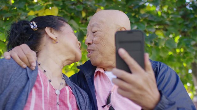 Elderly Asian couple pose for a selfie on their cell phone and share a kiss, in slow motion
