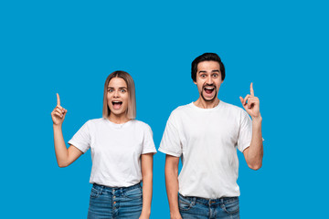 Smiling couple in white t-shirts and jeans pointing up with fingers standing isolated over blue background. Copy space for product advertisement.