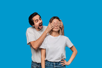 Brunet bearded man with mustaches in white t-shirts and blue jeans making a gift for a young blond woman isolated over blue background. Surprise for a girlfriend.