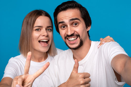 Happy couple of a young blond woman and brunet bearded man with mustaches in white t-shirts and blue jeans making selfie holding thumbs up isolated over blue background. Concept of an ideal couple.