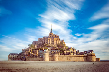 Fototapeta premium Mont Saint Michel and its abbey with the sea at low tide and clouds moving by the strong wind