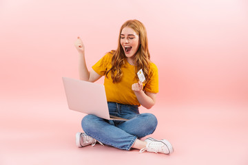 Happy girl isolated over pink wall background using laptop computer holding credit card make winner gesture.