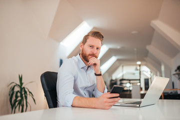 Portait of young redhead entrepreneur using smartphone and laptop at work.