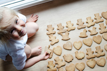 Smiling little girl makes a handmade cookies in different form on the wooden brown table in the kitchen. Top view.