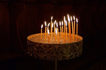 Light from lit candles in a candelabra stand filled with sand and soil in a Greek orthodox byzantine church interior at a monastery in Greece on dark background